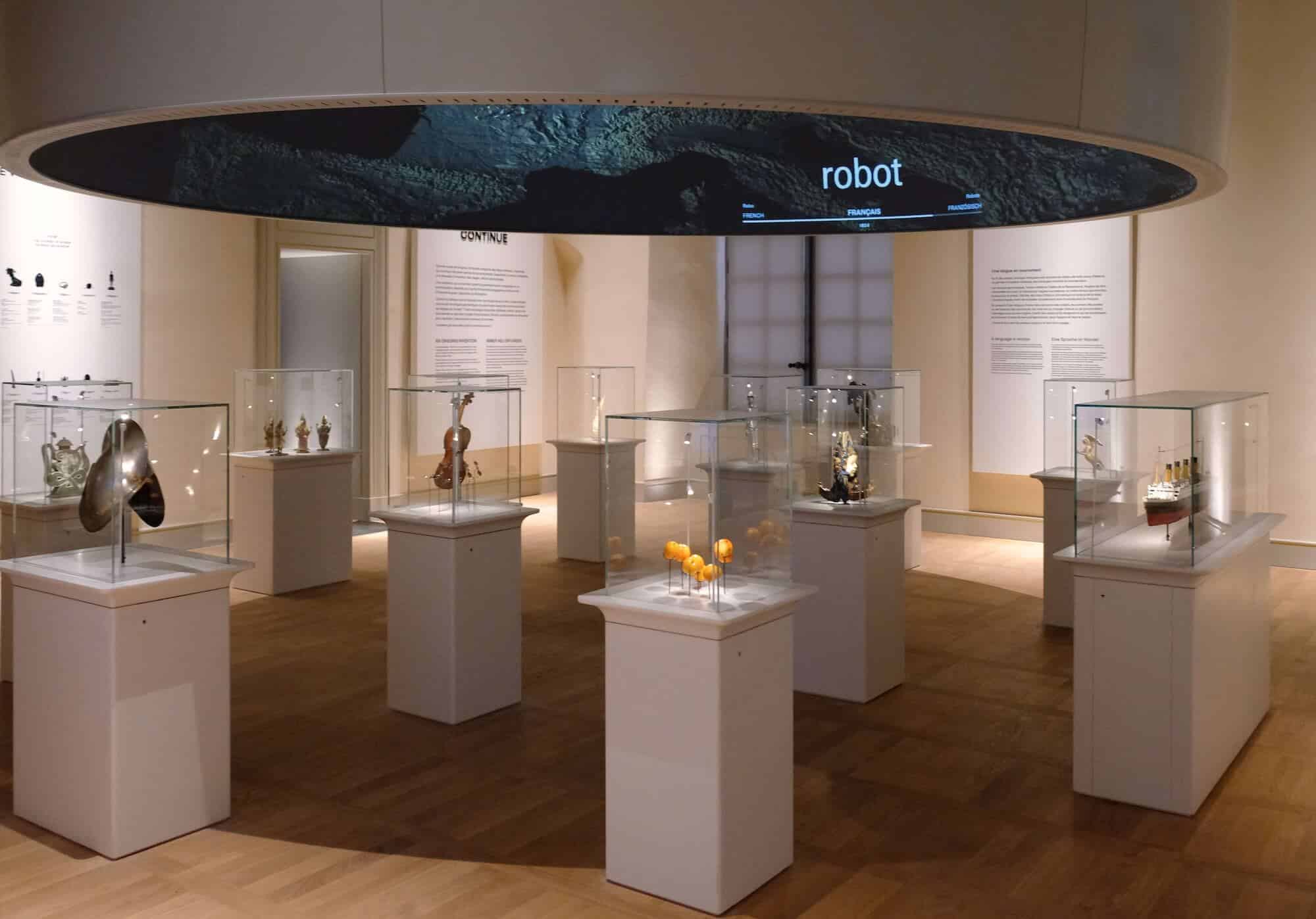 A circular suspended ring screen projecting the word 'robot' above museum display cases