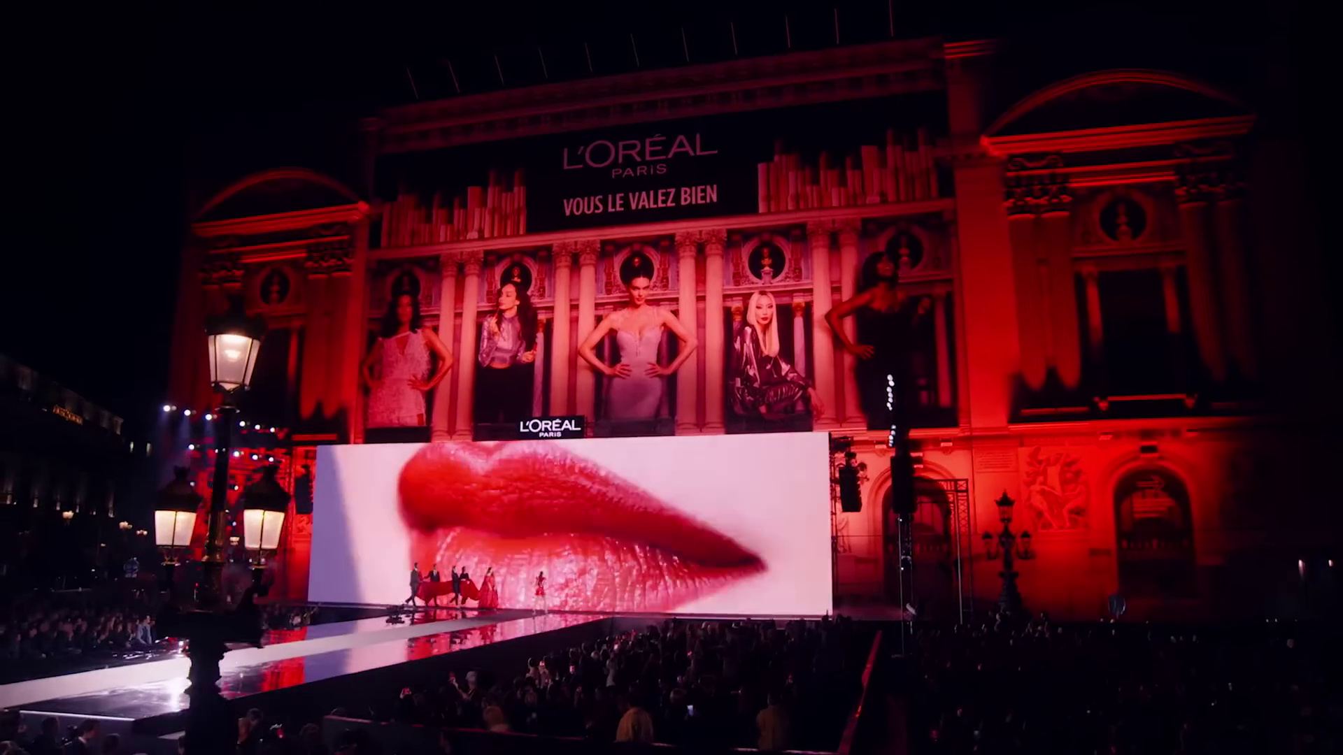 A giant LED screen of red lips on the Place de l'Opéra at night, the Opéra Garnier behind it