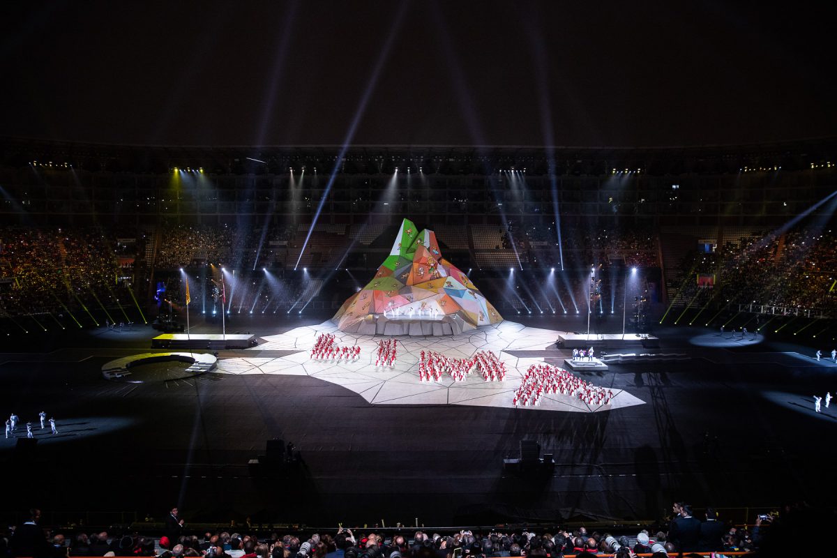 A vast stadium ceremony with performers forming the word LIMA before a giant mapped mountain set